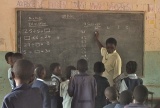 Children holding mini-blackboards with some sums on are stood in front of the main blackboard and teacher, who is looking to them for answers